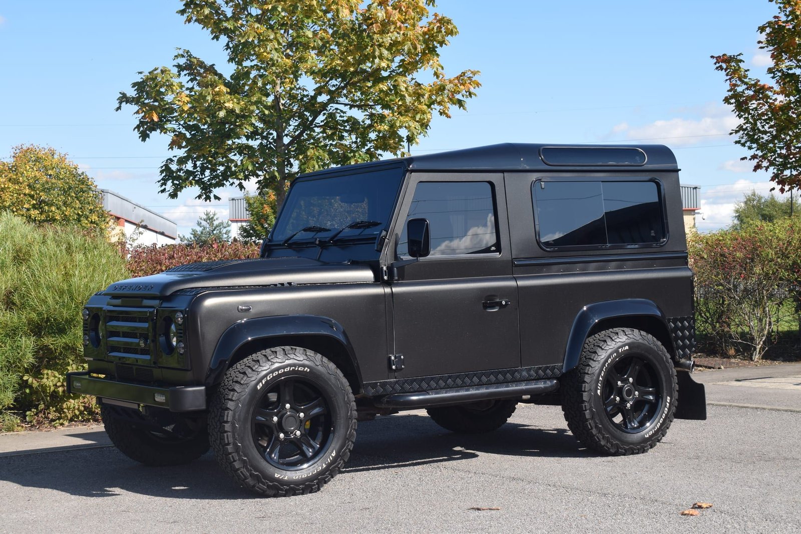 Black Land Rover Defender 90 with matte wrap finish, parked outdoors against trees and a blue sky.